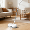 A white and black floor steam cleaner with a long handle stands on a light wood floor in a bright, modern living room with a beige sofa, wooden coffee table, and neutral rug.